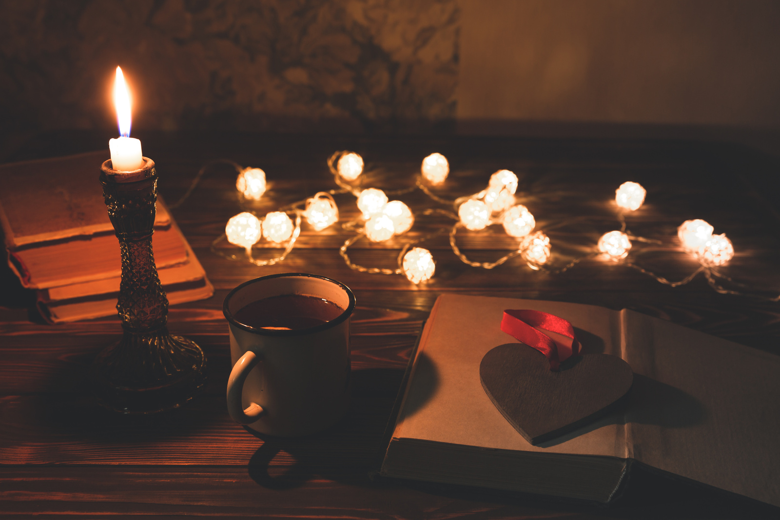 Book and hot tea mug for cosy evening. Holidays mood photo with christmas lights. Wooden heart and candle. Hygge concept. Perfect autumn flat lay.